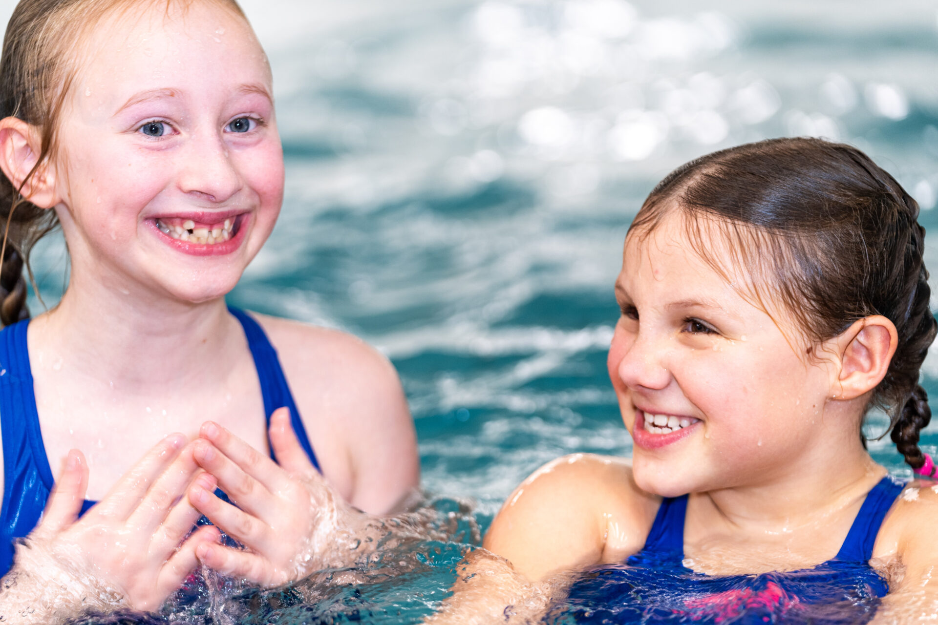 Zwei junge Mädchen in blauen Badeanzügen lächeln und lachen zusammen, während sie in einem Schwimmbecken spielen. Das Wasser plätschert um sie herum, und sie wirken glücklich und fröhlich.