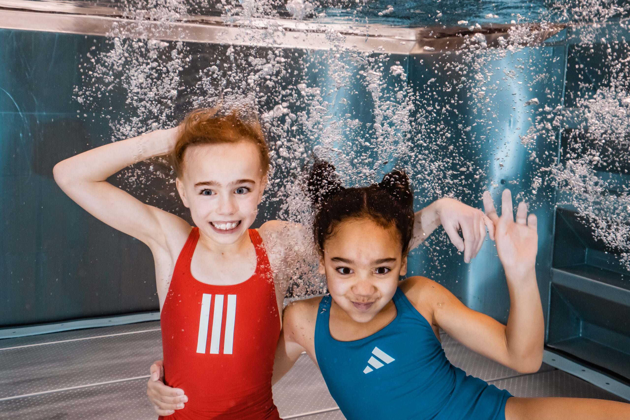 Zwei junge Mädchen in farbenfrohen Badeanzügen posieren und lächeln unter Wasser in einem Schwimmbad, umgeben von Blasen und hellem blauen Licht.