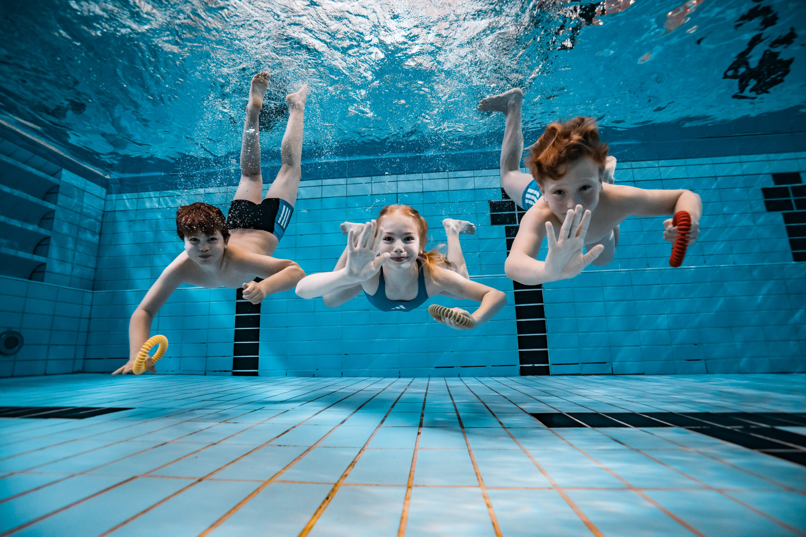 Drei Kinder schwimmen in einem Pool unter Wasser, lächeln und winken in die Kamera. Sie halten bunte Tauchspielzeuge und tragen Badeanzüge. Die Fliesen des Schwimmbeckens sind unter ihnen und um sie herum zu sehen.