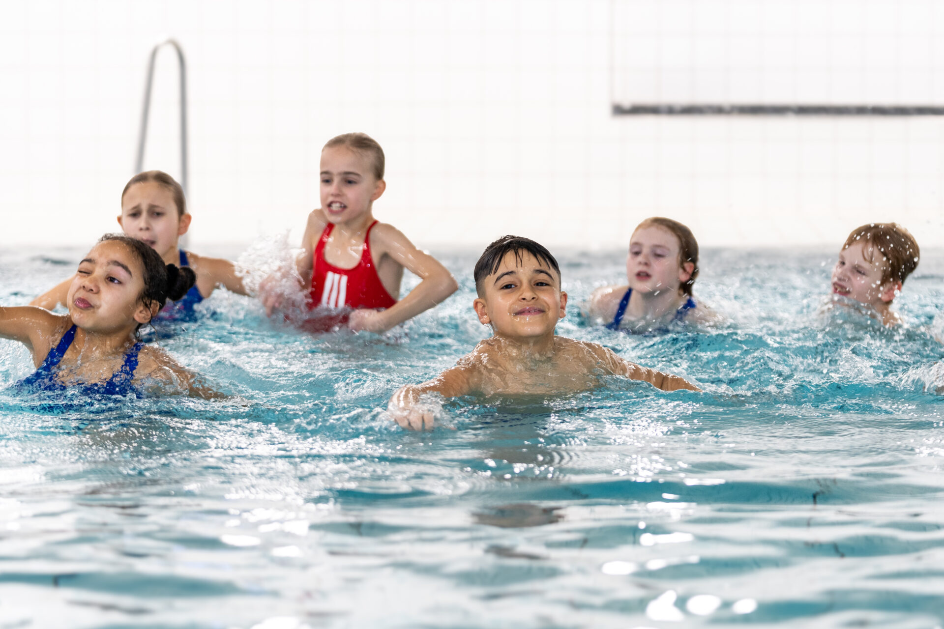 Eine Gruppe von sechs Kindern schwimmt und spielt zusammen in einem Hallenbad, spritzt Wasser und lächelt. Der Hintergrund ist eine weiß gekachelte Wand.