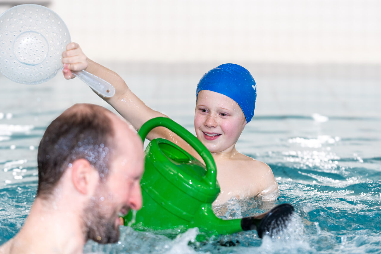 Ein lächelndes Kind mit blauer Badekappe spielt in einem Swimmingpool und hält eine grüne Gießkanne und ein weißes Sieb in der Hand, während im Vordergrund ein Erwachsener mit rasiertem Kopf zu sehen ist.