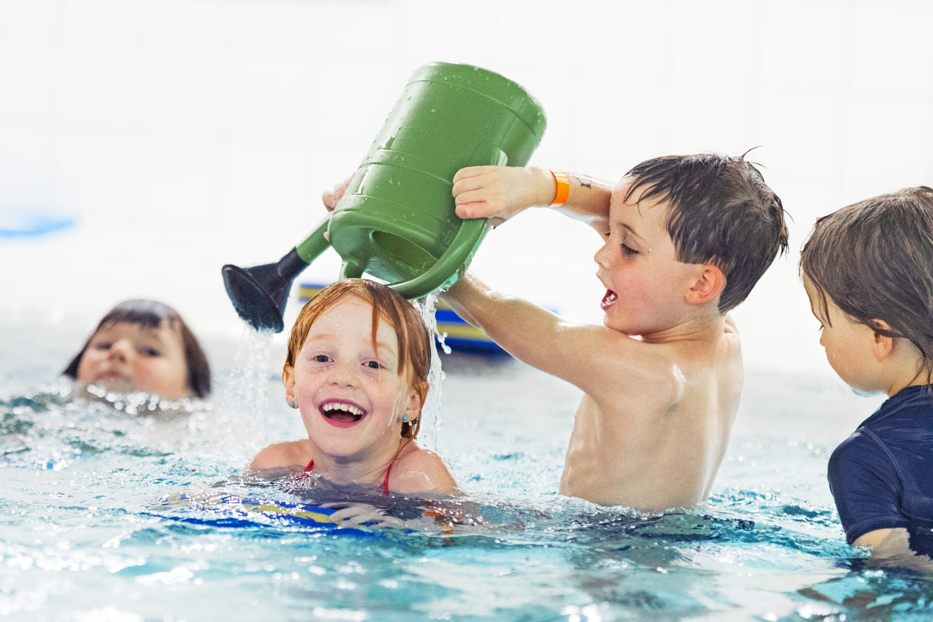 Vier Kinder spielen in einem Schwimmbecken. Ein Junge schüttet Wasser aus einer großen grünen Gießkanne über ein lächelndes rothaariges Mädchen, während zwei andere Kinder in der Nähe zusehen und lachen.
