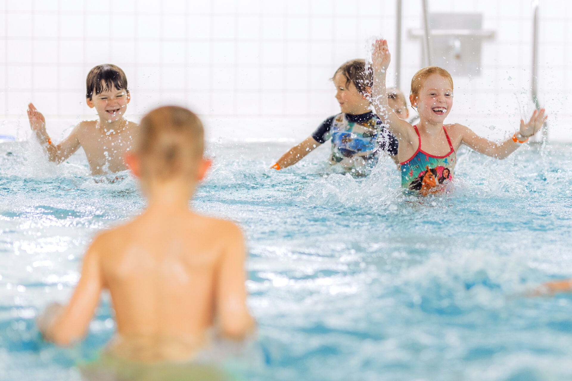 Vier Kinder spielen und planschen in einem hell erleuchteten Hallenbad, lächeln und heben ihre Arme. Das Bild ist fröhlich und energiegeladen mit gekachelten Wänden im Hintergrund.
