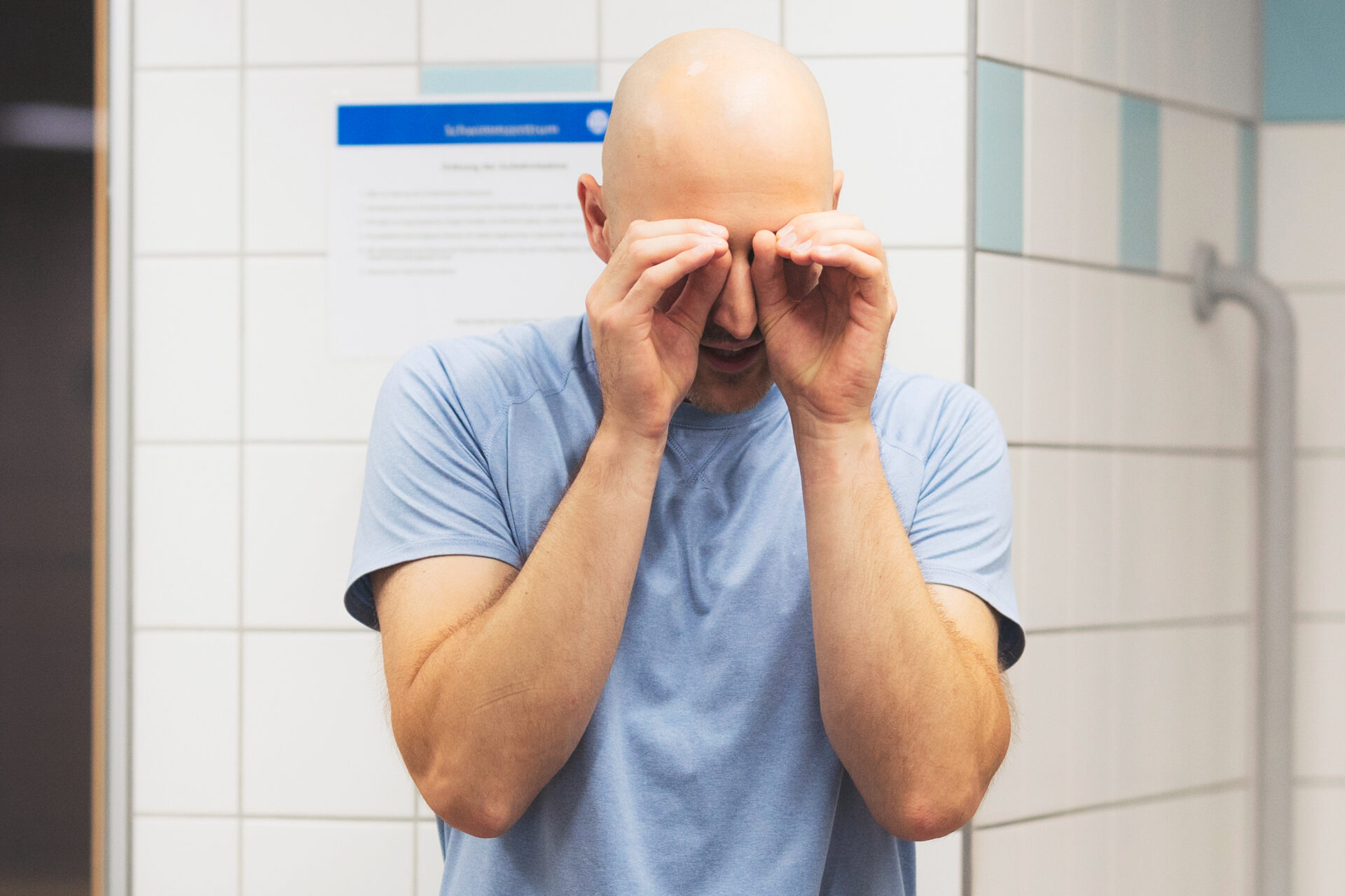 Ein glatzköpfiger Mann in einem hellblauen Hemd steht in einem gekachelten Raum und reibt sich mit beiden Händen die Augen. An der gekachelten Wand hinter ihm ist ein verschwommenes Schild zu sehen.