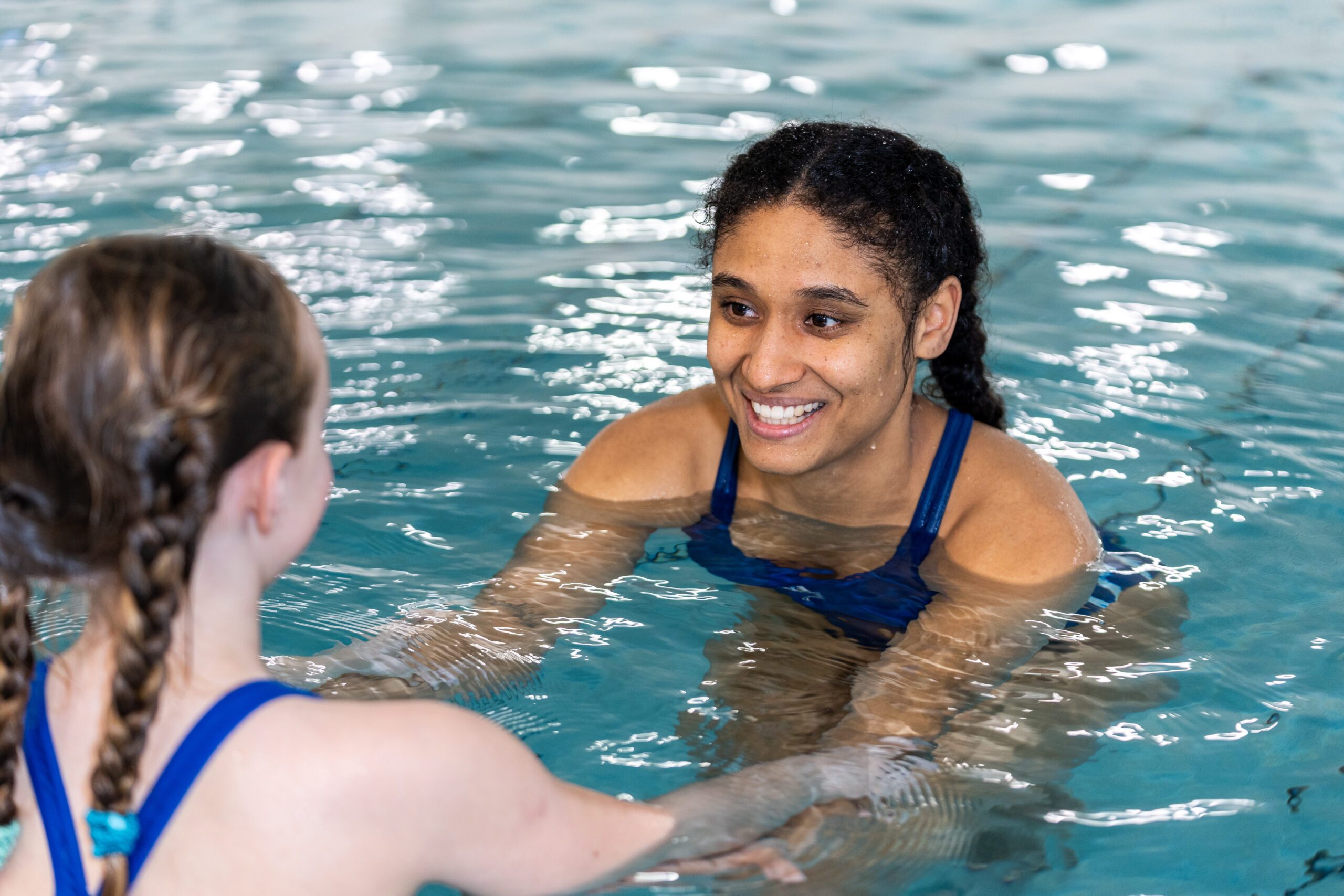 Eine lächelnde Schwimmlehrerin in einem blauen Badeanzug hält einem jungen Mädchen im Schwimmbecken die Hand und gibt ihm während einer Schwimmstunde Anweisungen.