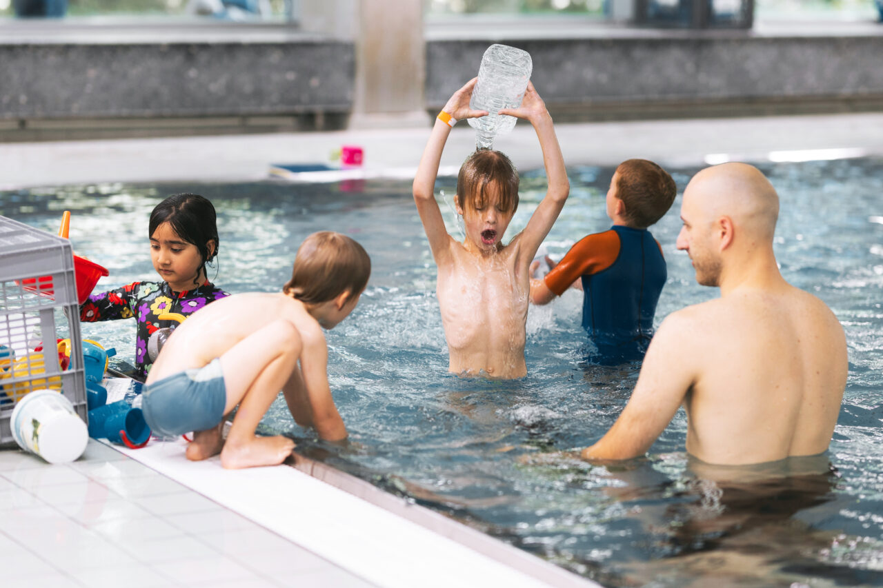 Vier Kinder und ein Erwachsener spielen in einem Hallenbad. Ein Kind schüttet sich Wasser aus einer Flasche über den Kopf, während die anderen mit Poolspielzeug beschäftigt sind oder im Wasser interagieren.
