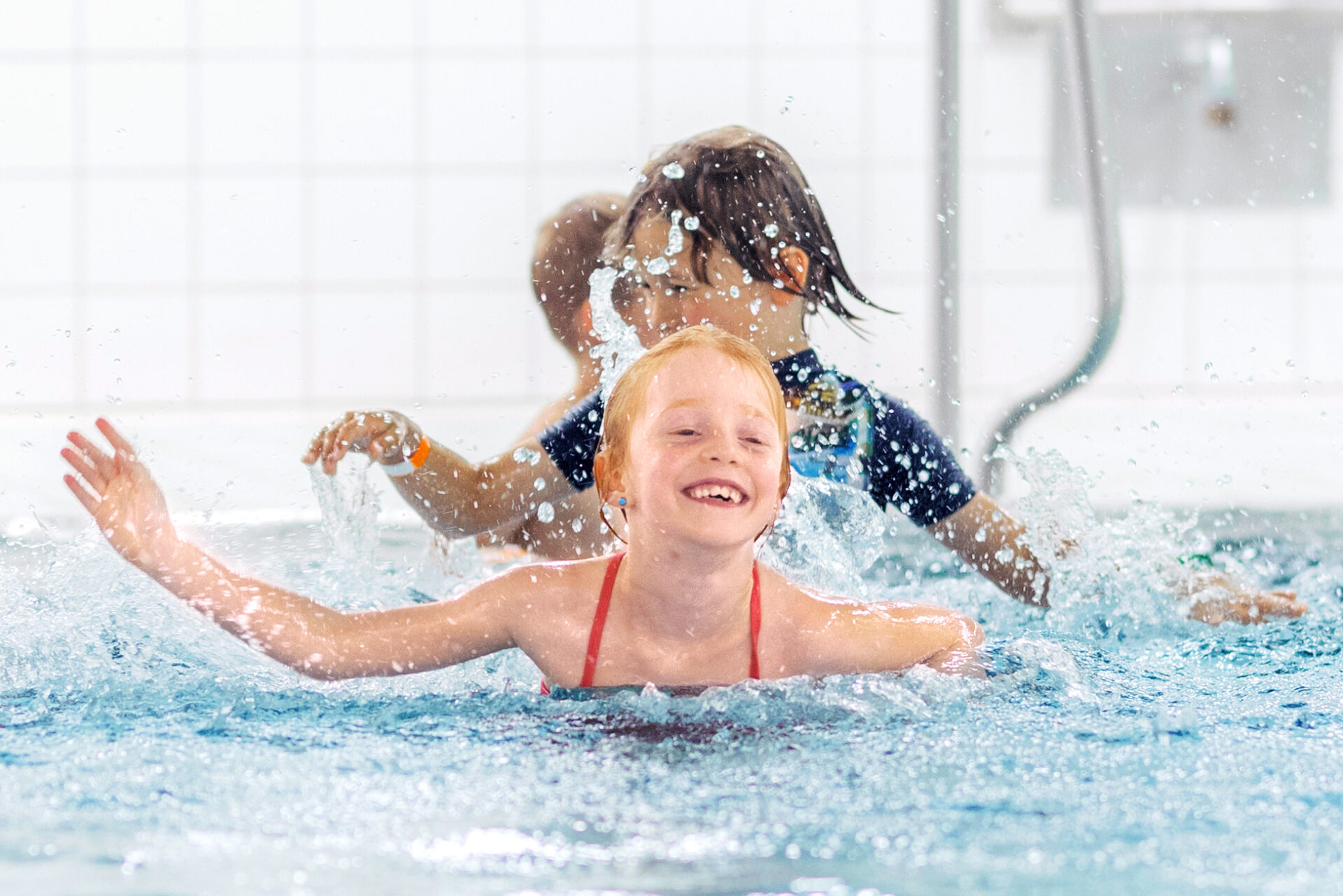 Zwei Kinder spielen und planschen in einem Schwimmbecken. Das Kind im Vordergrund lächelt fröhlich, während ein anderes Kind hinter ihnen mit Wasser bespritzt wird. Der Hintergrund zeigt eine geflieste Wand und eine Badeleiter.
