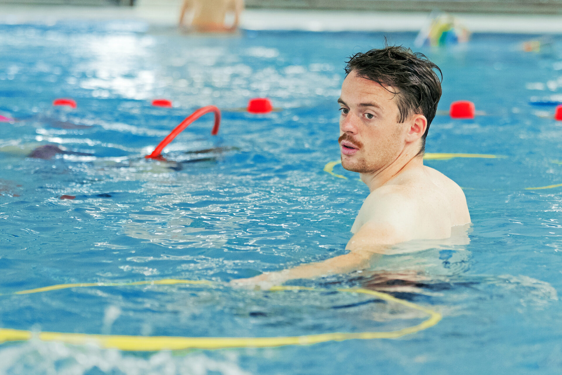 Ein Mann mit nassen Haaren steht in einem Schwimmbecken und schaut über seine Schulter in Richtung Kamera. Um ihn herum schwimmen gelbe Reifen und rote Markierungen auf dem Wasser.