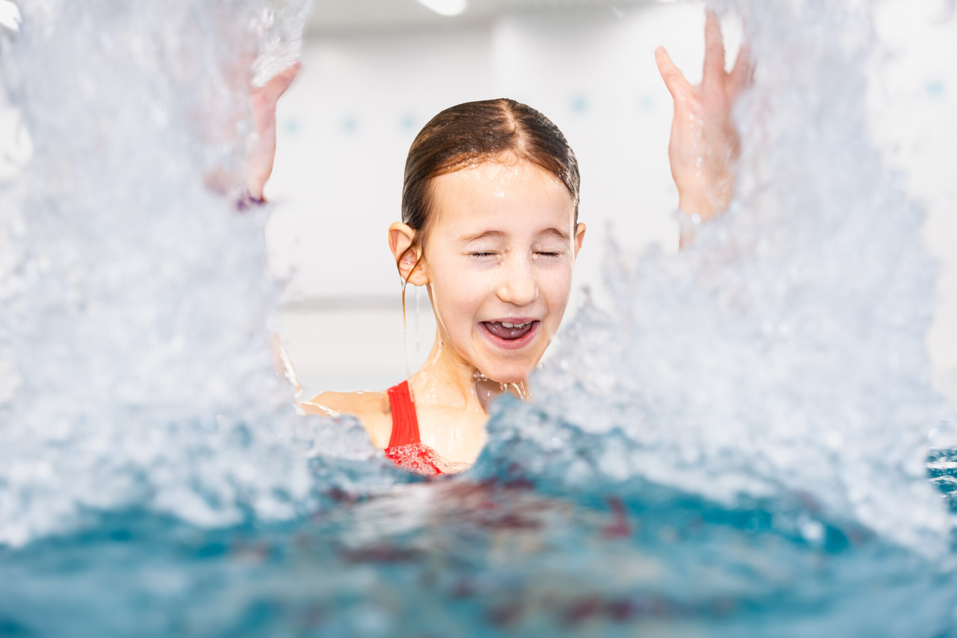 Ein junges Mädchen in einem roten Badeanzug spritzt mit geschlossenen Augen lächelnd im Wasser, während das Wasser um sie herum in einem Schwimmbad spritzt.