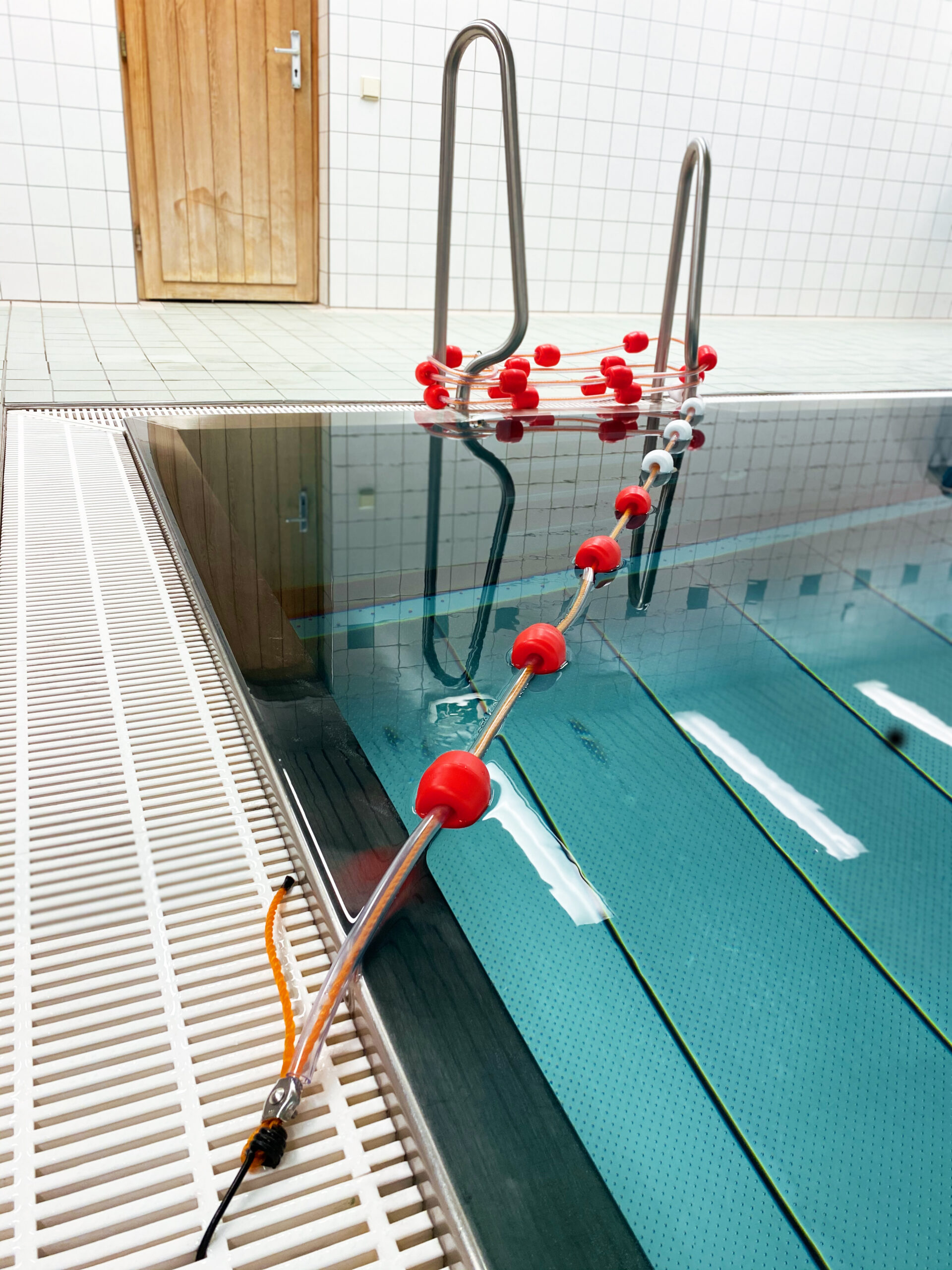 Ein Hallenbad mit einer Edelstahlleiter, einer Bahntrennung mit roten Schwimmern und einer Holztür im Hintergrund. Der Poolbereich hat weiß geflieste Wände und ein Abflussgitter am Rand.