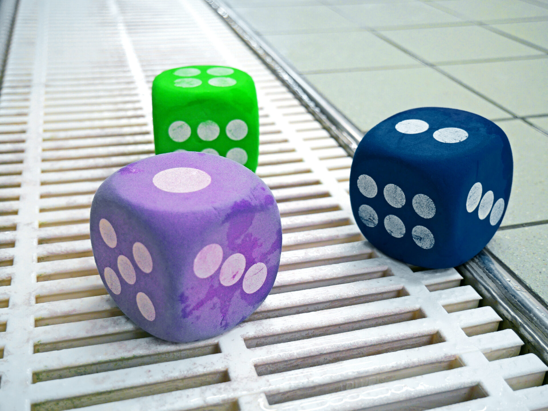 Drei große Schaumstoffwürfel - ein grüner, ein blauer und ein lila Würfel - liegen auf einem weißen Gitter neben beigen Bodenfliesen. Jeder Würfel zeigt eine andere Anzahl von weißen Punkten.